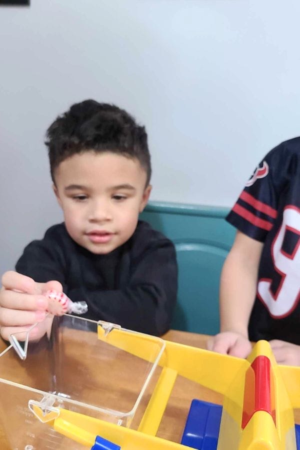 A young child is playing a tabletop game, holding a small game piece above a plastic game board—perfect for inspiring fun Christmas Math Activities. Another child wearing a sports jersey is partially visible next to him.