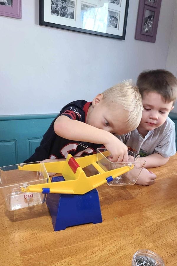 Two young boys sit at a table, intently playing with a yellow and blue balance scale toy, placing objects in the trays to compare their weights as part of fun Christmas Math Activities.