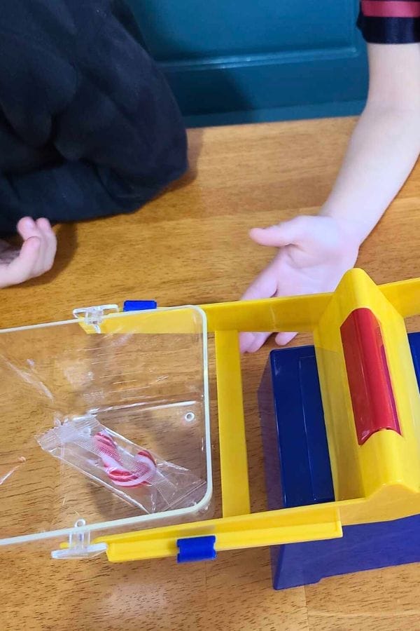 A child's hands near a yellow and blue plastic balance scale with a wrapped peppermint candy on one side, on a wooden table—perfect for fun Christmas Math Activities.