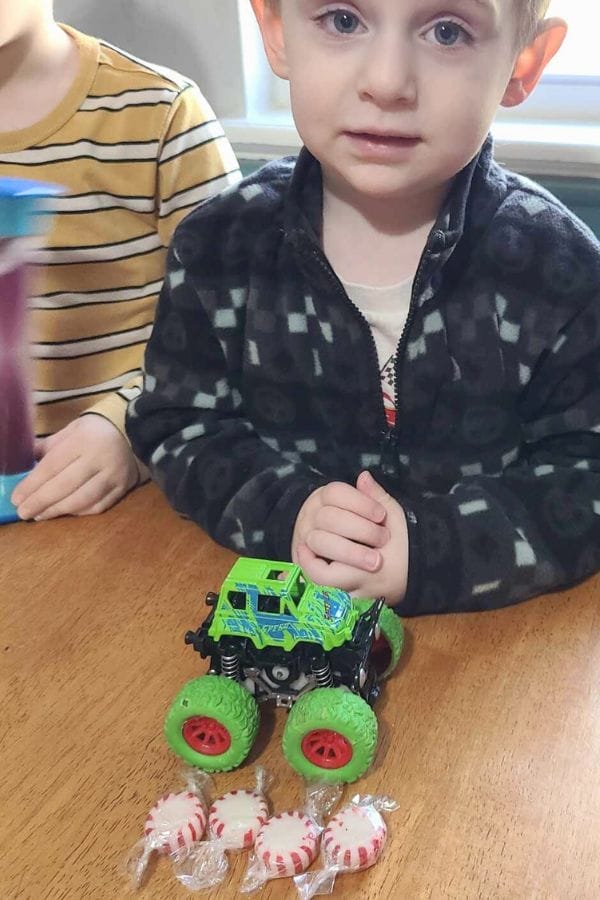A young child sits at a table with a green toy monster truck and five peppermint candies laid out in front of them, exploring Christmas Math Activities. Another child is partially visible to the left.