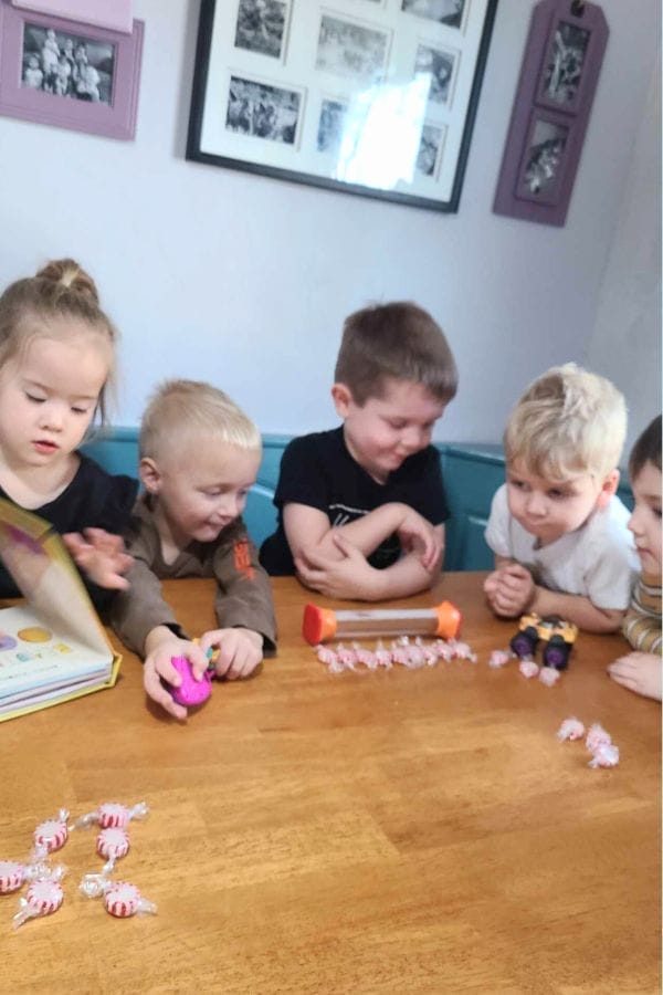 Five young children sit at a table with candies and toys, engaging in Christmas Math Activities as they look at objects in front of them. Framed photos hang on the wall in the background.