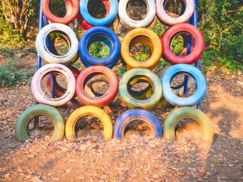 A playground climbing structure made of colorful tires stacked in a grid, crafted from DIY Outdoor Play Materials, with sunlight and dry leaves on the ground nearby.