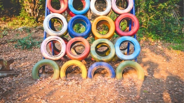 A playground climbing structure made of colorful tires stacked in a grid, crafted from DIY Outdoor Play Materials, with sunlight and dry leaves on the ground nearby.