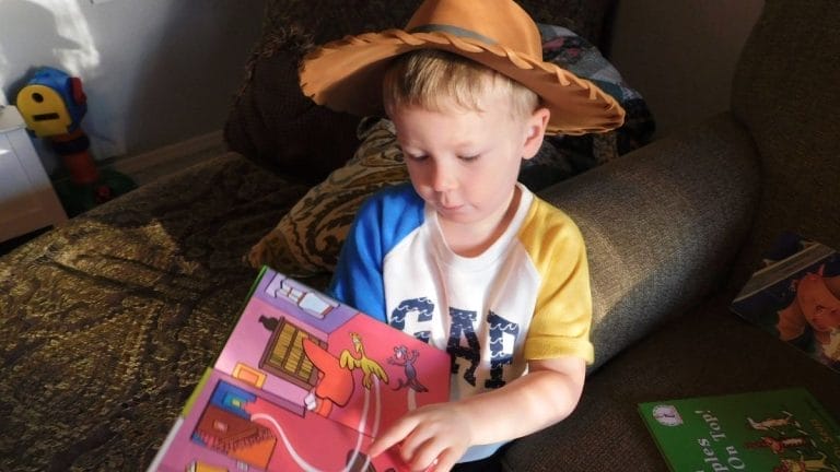 Young child wearing a brown cowboy hat sits on a couch reading a colorful picture book, with more books nearby, perhaps inspired by Lesson Plans on The Cat in the Hat.