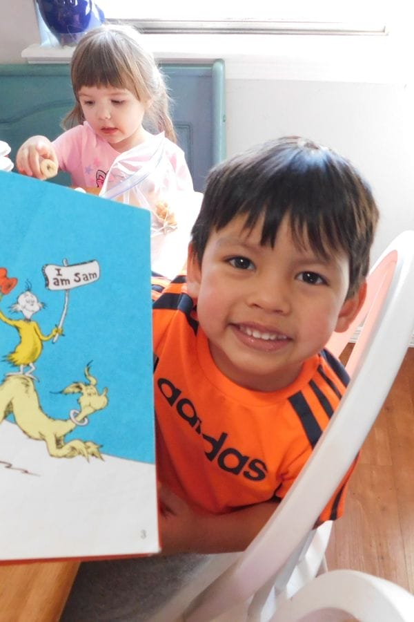 A young boy in an orange shirt smiles while holding a Dr. Seuss book open to a page that says "I am Sam"; he’s excited for lesson plans on The Cat in the Hat, while a young girl sits at the table behind him.