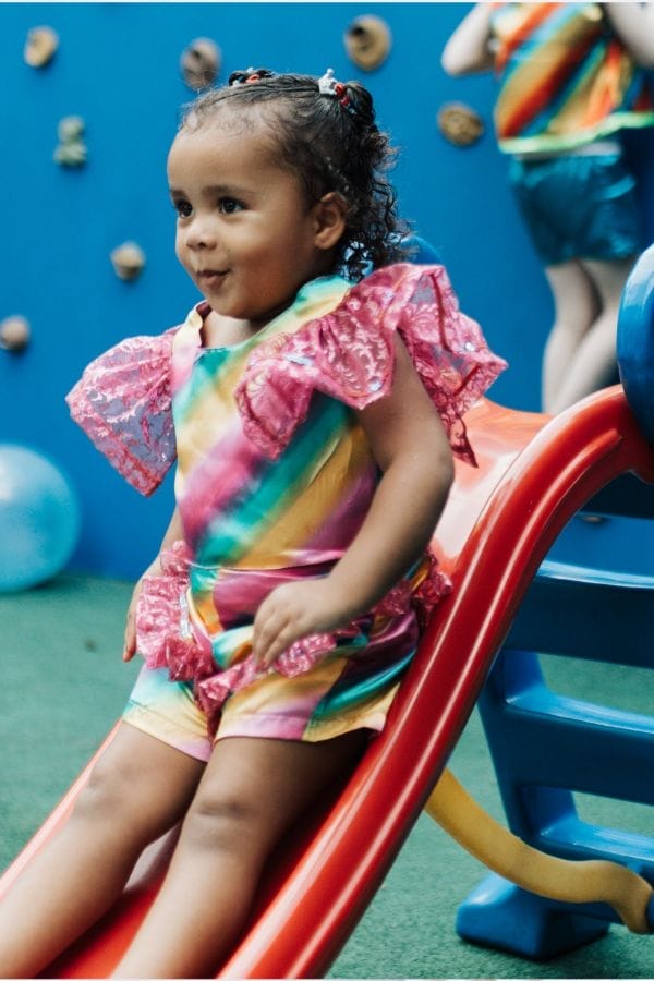 A young child in a colorful outfit sits at the top of a red slide, with a blue climbing wall and another child in the background—perfect for showcasing how Growing Your Daycare Business creates vibrant play spaces for kids.