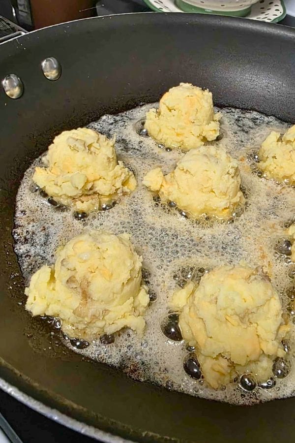 Six scoops of dough, shaped into Mashed Potato Cakes, are being deep-fried in bubbling oil in a large black pan.