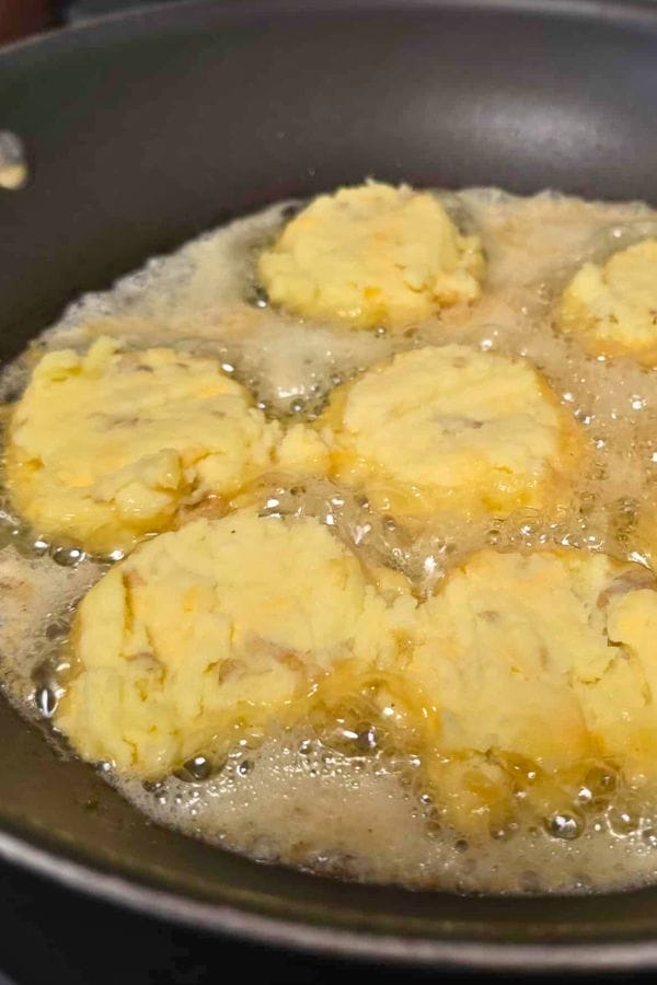 Golden Mashed Potato Cakes are being deep fried in hot oil in a pan, forming crispy, round patties.