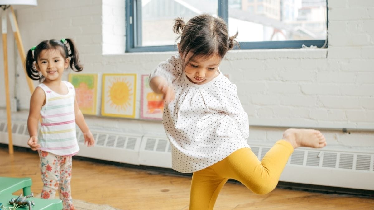 Two young girls play indoors at a New Year’s Eve Kid’s Party; one kicks her leg up while the other stands smiling in the background. Bright artwork hangs on the white brick wall behind them.