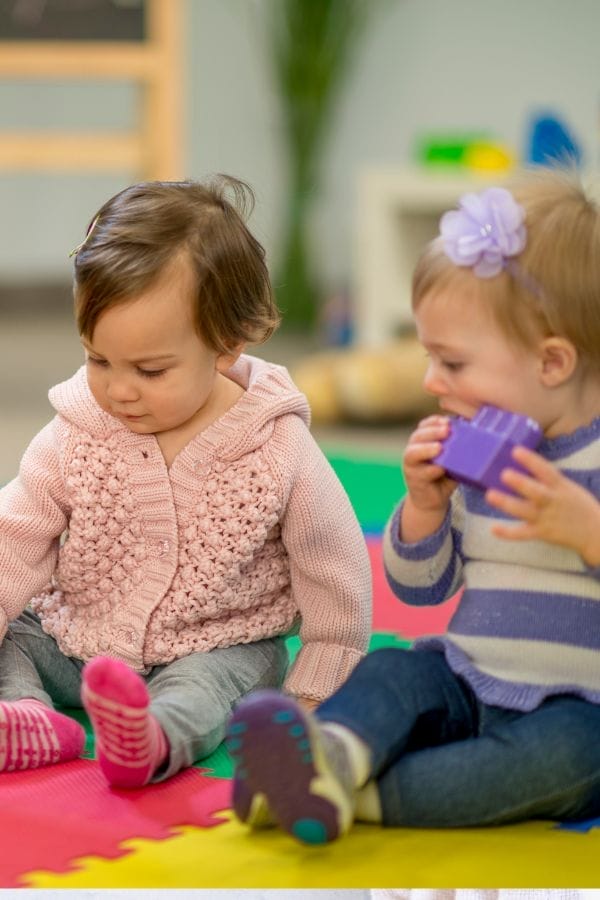 Two toddlers sit on a colorful foam mat at a New Year's Eve Kid’s Party; one looks down at the floor while the other holds and examines a purple toy block.