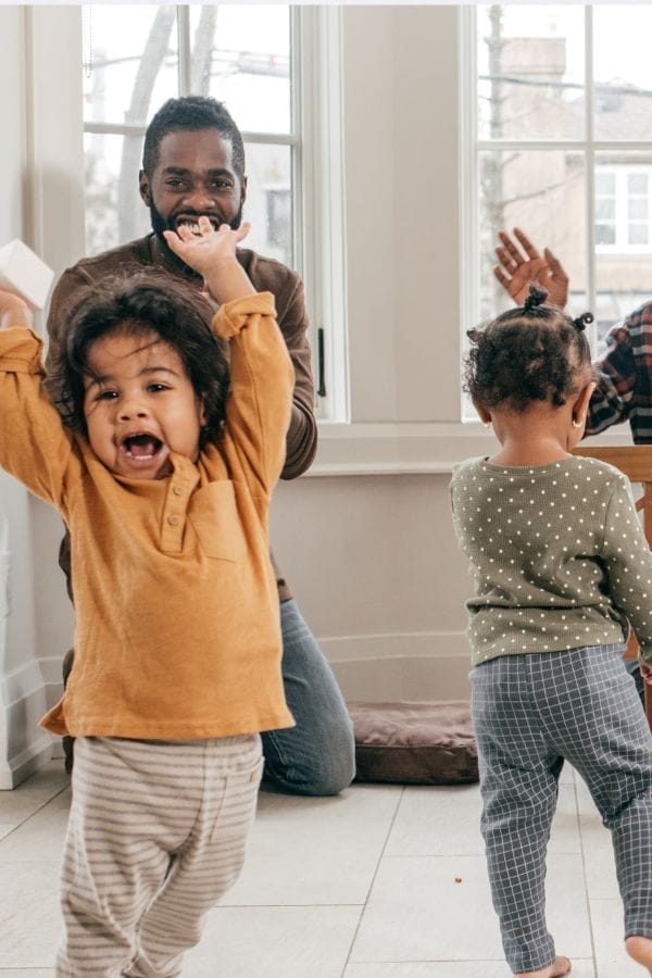 A man sits in a bright room smiling while two young children play nearby at a New Year’s Eve Kid’s Party; one child faces the camera with arms raised, and the other stands with their back turned.