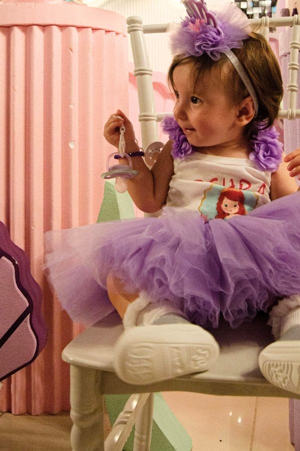 A young child in a purple tutu and headband sits on a white chair, holding a pacifier and looking to the side at a New Year’s Eve Kid’s Party, with festive pink and purple decorations in the background.