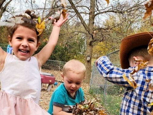 Three young children play with fallen leaves outside; one girl smiles with arms raised, another boy wears a cowboy hat, and a third child sits nearby. This joyful scene is perfect inspiration for themed play based lesson plans. Trees and playground equipment are in the background.