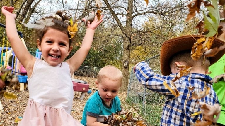 Three young children play with fallen leaves outside; one girl smiles with arms raised, another boy wears a cowboy hat, and a third child sits nearby. This joyful scene is perfect inspiration for themed play based lesson plans. Trees and playground equipment are in the background.