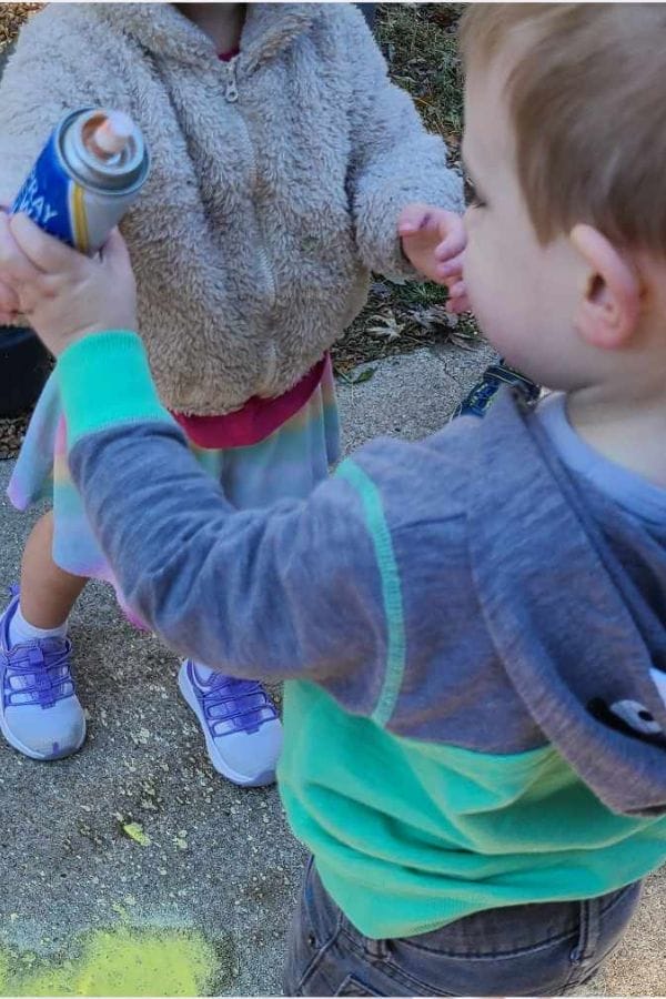 Two young children are outdoors; one child holds an aerosol spray can while the other reaches out. Yellow spray paint is visible on the pavement, hinting at creative fun inspired by Themed Play Based Lesson Plans.