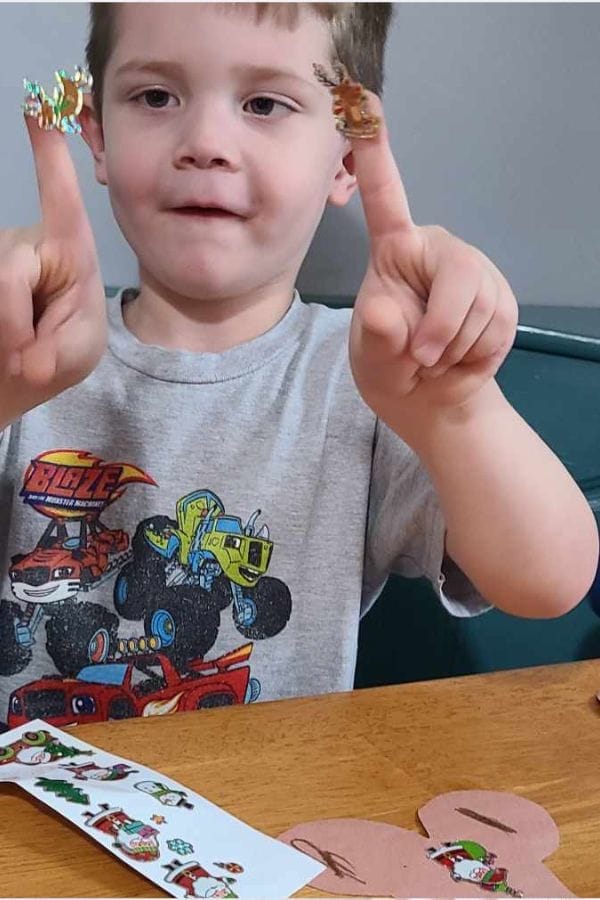 A young boy wearing a monster truck t-shirt holds up his index fingers, each with a holiday-themed sticker on it, while sitting at a table with more stickers and paper—perfect inspiration for themed play based lesson plans.