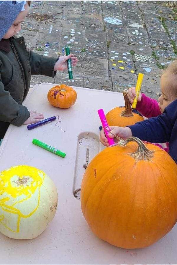 Two children use colored markers to decorate pumpkins on a table outdoors, enjoying hands-on creativity inspired by themed play based lesson plans.