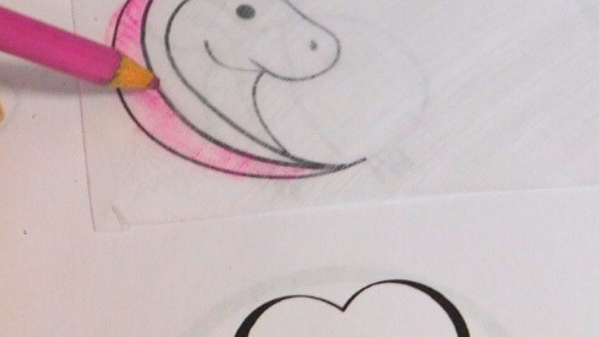 A pencil colors a pink outline on tracing paper over a simple cartoon drawing of a smiling dinosaur head inside a heart shape, perfect for creating Superhero Shrinky Dink Patterns.