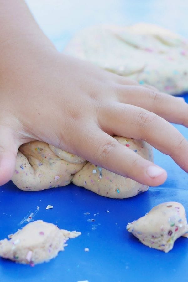 A child's hand presses into a lump of homemade, eco friendly craft play dough with colorful specks on a blue surface.