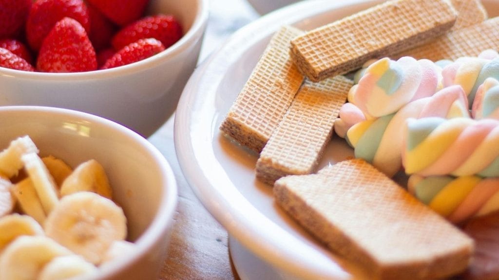 Bowls of sliced bananas and strawberries sit next to a plate with wafer cookies and pastel-colored twisted marshmallows—perfect New Year’s Eve food for a festive celebration.