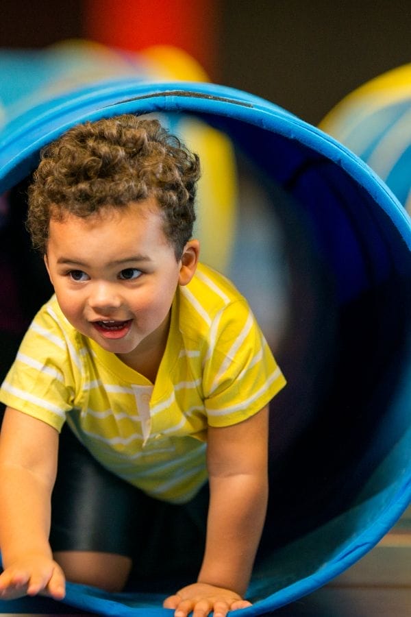 A young child in a yellow striped shirt crawls through a blue play tunnel, smiling and looking ahead—one of many fun STEM activities for toddlers that encourage exploration and learning.