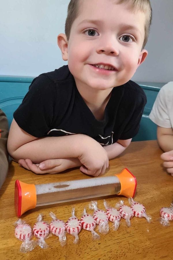 A young boy sits at a table and smiles at the camera, with a row of wrapped peppermint candies and a sand timer in front of him—perfect for fun STEM Activities for Toddlers.