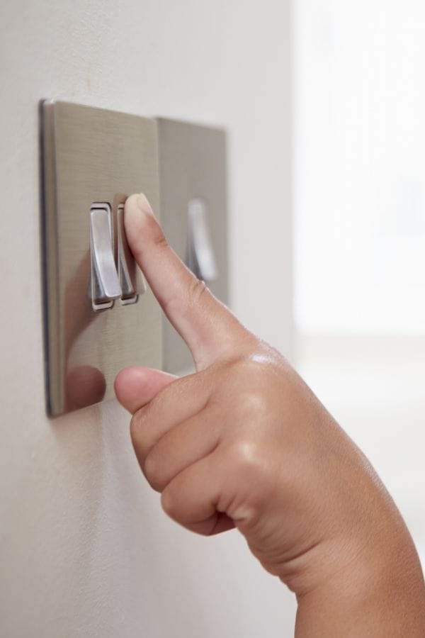 A child's hand is flipping a light switch on a wall-mounted switch plate, illustrating sustainable living with kids by teaching them energy-saving habits early.