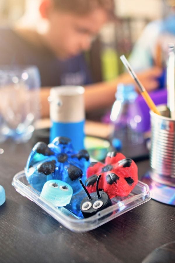 Plastic container holding painted egg carton insect crafts, perfect for teaching kids about recycling, with a blurred child and craft supplies in the background.