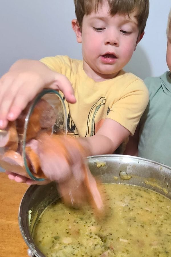 A young child explores Preschool Recipes by dropping sliced sausage from a glass bowl into a pot of thick, chunky soup while sitting at the table.