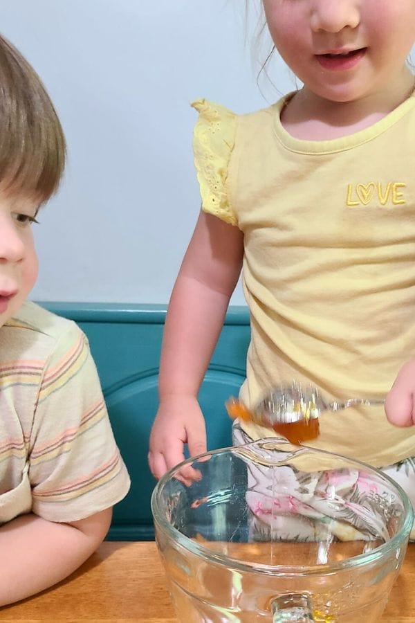 Two young children, exploring Preschool Recipes, prepare ingredients at a table—one carefully holds a spoon with honey over a glass mixing bowl.