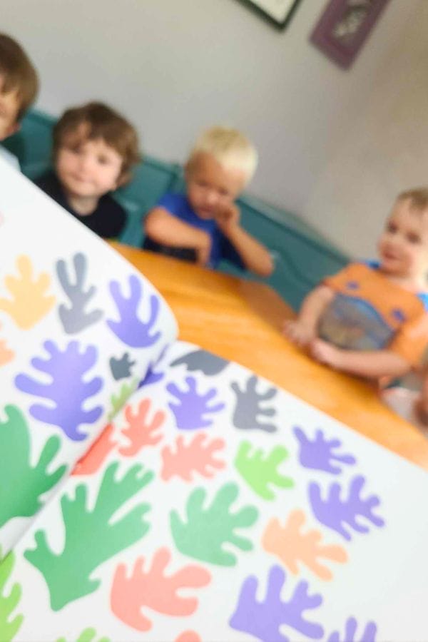 Four young children sit at a table in the background while an open book with colorful abstract shapes, perfect for inspiring lesson plans for toddlers, is in focus in the foreground.