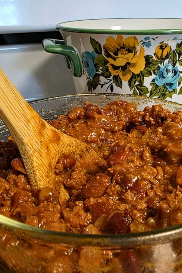 A wooden spoon stirs a pot of chili with beans and ground meat; a floral-patterned pot sits in the background on the stove.