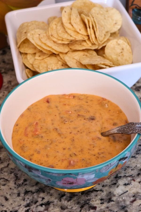 A bowl of cheesy dip with a spoon next to a square dish filled with tortilla chips on a granite countertop.