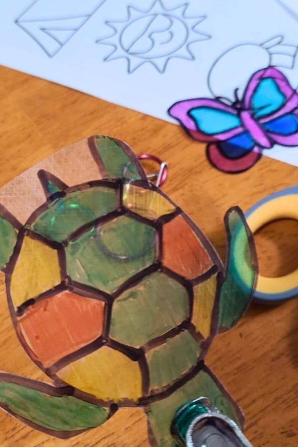 A colored turtle-shaped keychain made from Spring Shrinky Dink Patterns is in the foreground, with a partially colored butterfly drawing and washi tape on a wooden table in the background.