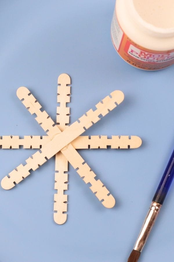 Six interlocking wooden craft sticks arranged in a star shape on a blue surface, with a paintbrush and Mod Podge nearby—perfect for Snowflake Crafts for Kids.