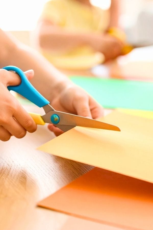 Close-up of a person using blue-handled scissors to cut yellow construction paper on a table with other colored papers—perfect for making Snowflake Crafts for Kids.