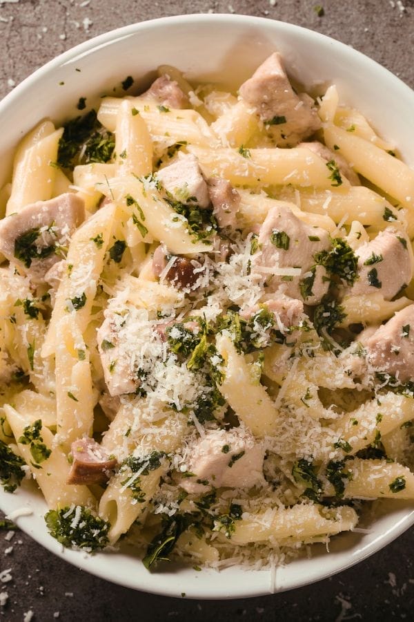 A bowl of penne pasta mixed with leftover chicken breast, chopped herbs, and grated Parmesan cheese.
