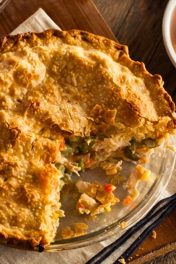 A baked chicken pot pie with a golden, flaky crust in a glass pie dish, missing a few slices to reveal a filling of vegetables and leftover chicken breast.