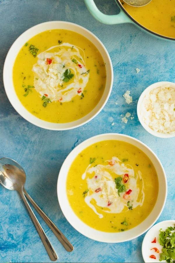 Two bowls of yellow soup, inspired by healthy soup recipes, are garnished with cream, herbs, and red chili slices on a blue surface, accompanied by spoons and a small bowl of white flakes.