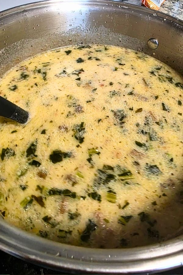 A pot of creamy Loaded Zuppa Toscana Soup with leafy greens and visible herbs simmers on the stovetop, a black ladle partially submerged in the hearty mixture.