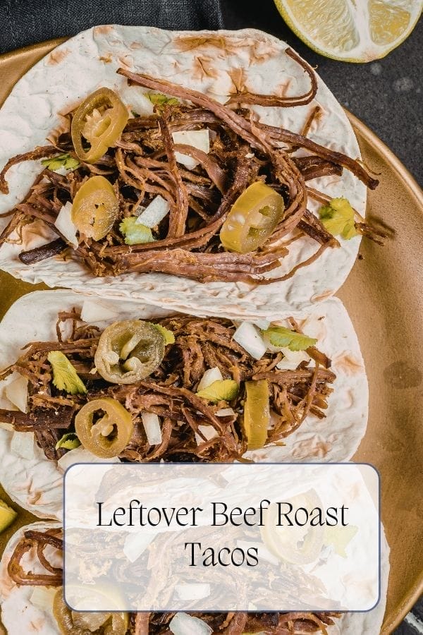 Two tacos filled with tender leftover beef roast, sliced jalapeños, chopped onions, and cilantro sit on a gold plate, with a halved lemon in the background. Text reads "Leftover Beef Roast Tacos.